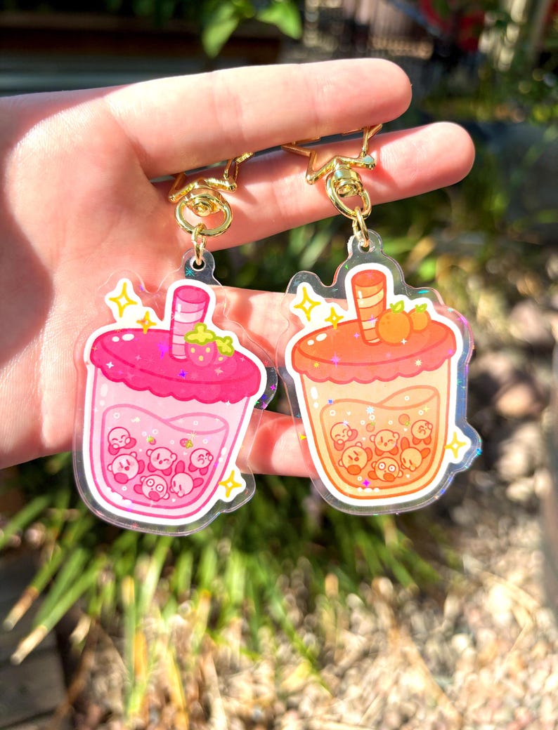 May include: Two acrylic keyrings with bubble tea designs. One is pink with a strawberry, the other is orange with oranges. Both keyrings have gold-coloured clasps and include cartoon characters inside the drinks.