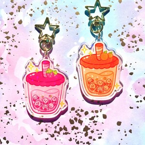 May include: Two acrylic keychains in the shape of bubble tea drinks. One is pink with a strawberry on top, the other is orange with two small orange fruits. Both have a star-shaped clasp and a gold-coloured ring.