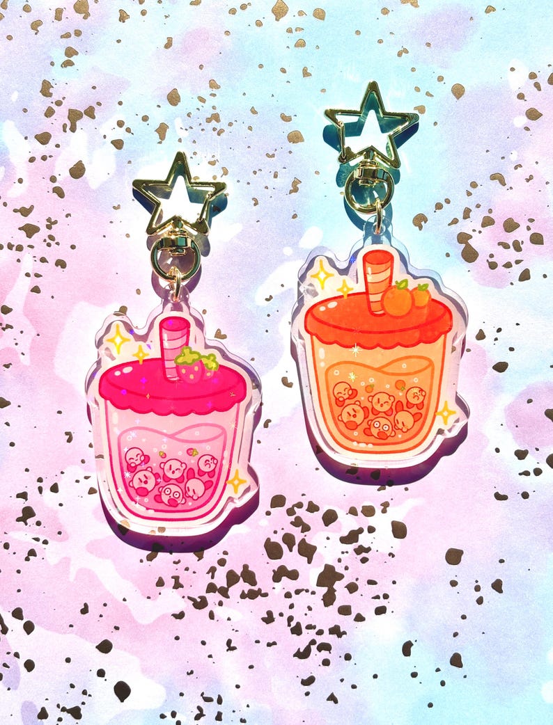 May include: Two acrylic keychains in the shape of bubble tea drinks. One is pink with a strawberry on top, the other is orange with two small orange fruits. Both have a star-shaped clasp and a gold-coloured ring.