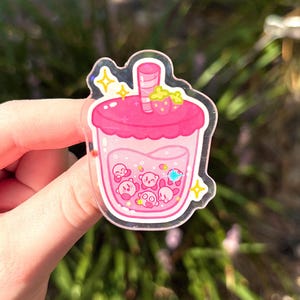 May include: A clear acrylic charm in the shape of a pink bubble tea. The charm features a pink drink with Kirby characters inside. A strawberry and yellow stars are on top. The charm is held by a hand.