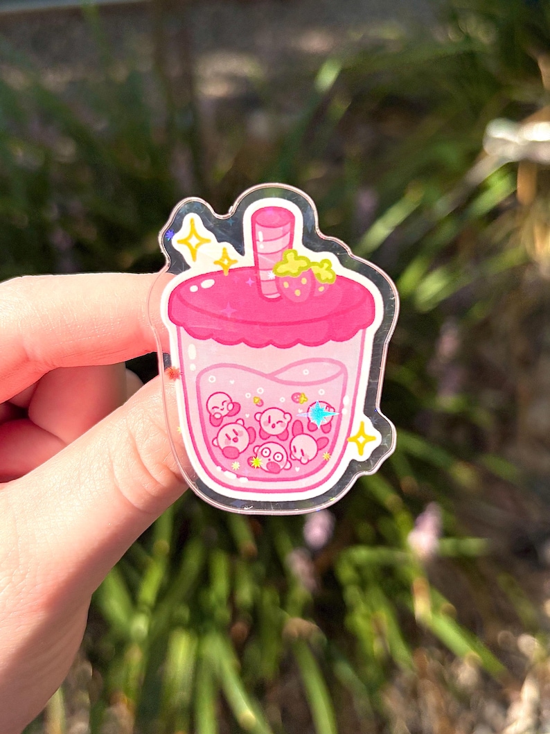 May include: A clear acrylic charm in the shape of a pink bubble tea. The charm features a pink drink with Kirby characters inside. A strawberry and yellow stars are on top. The charm is held by a hand.