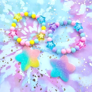 Può includere: Due braccialetti colorati con perline a forma di stella e un grande ciondolo a forma di stella. I braccialetti sono realizzati con perline rosa, gialle, blu e viola. I ciondoli sono iridescenti e hanno una finitura scintillante.