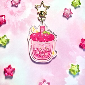 May include: A pink acrylic keyring depicting a cartoon drink with a strawberry garnish. Inside the drink are several small, round, pink characters. The keyring has a gold star-shaped clasp and is surrounded by star-shaped beads.