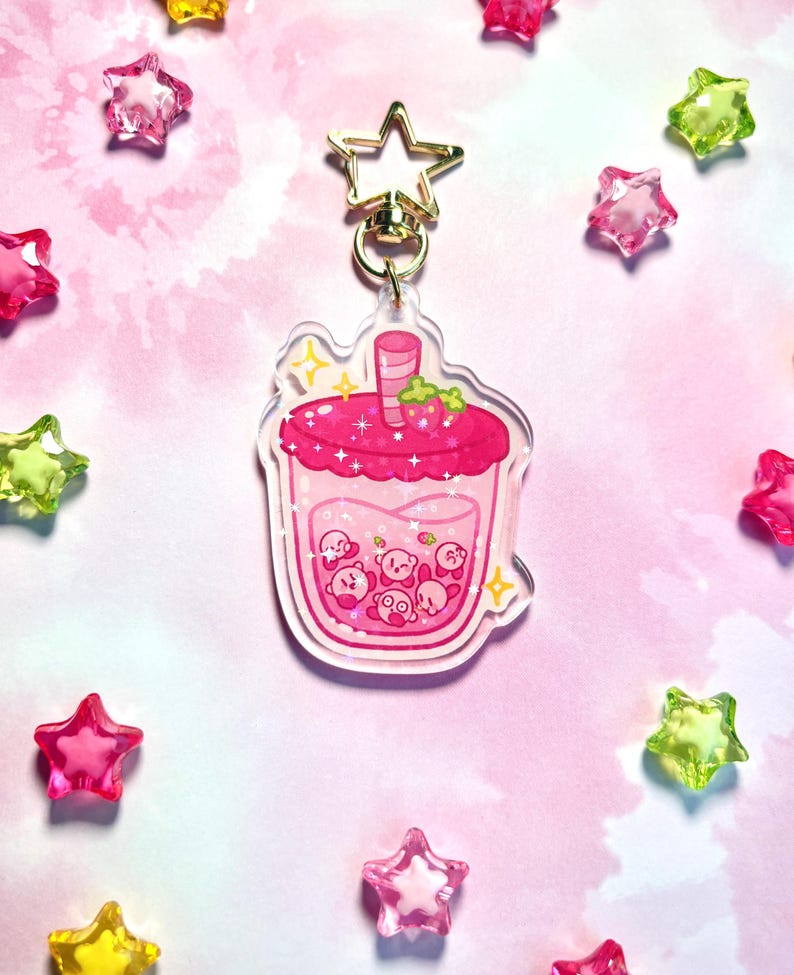 May include: A pink acrylic keyring depicting a cartoon drink with a strawberry garnish. Inside the drink are several small, round, pink characters. The keyring has a gold star-shaped clasp and is surrounded by star-shaped beads.