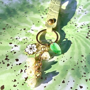 May include: A light green keychain with gold-colored hardware and a decorative charm cluster. The charms include a white flower, a green leaf, and a chain of small, cream-colored bell-shaped flowers. The keychain strap has a textured, crocodile-like pattern.