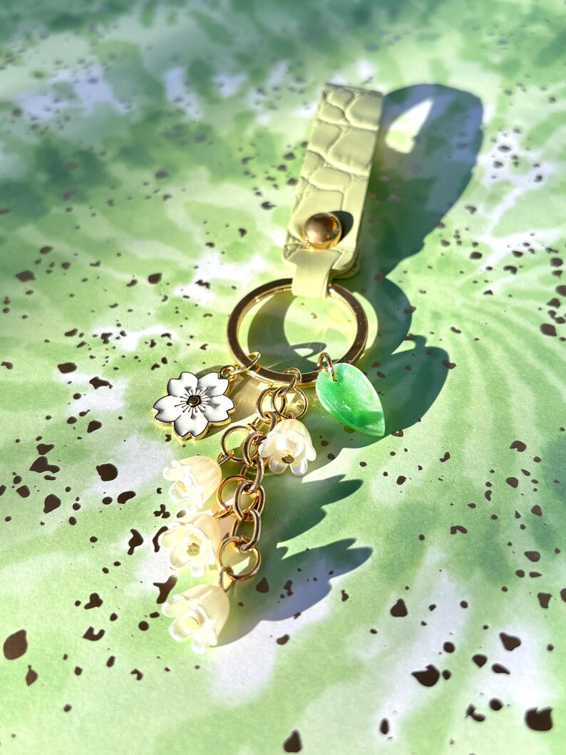 May include: A light green keychain with gold-colored hardware and a decorative charm cluster. The charms include a white flower, a green leaf, and a chain of small, cream-colored bell-shaped flowers. The keychain strap has a textured, crocodile-like pattern.