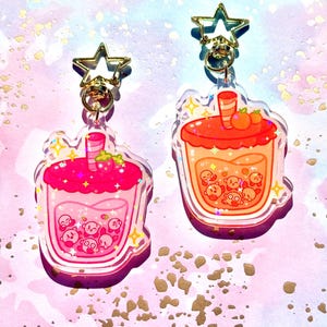 May include: Two acrylic keychains in the shape of bubble tea drinks. One is pink with a strawberry topping, the other is orange with a peach topping. Both have gold star-shaped clasps and feature cartoon characters.