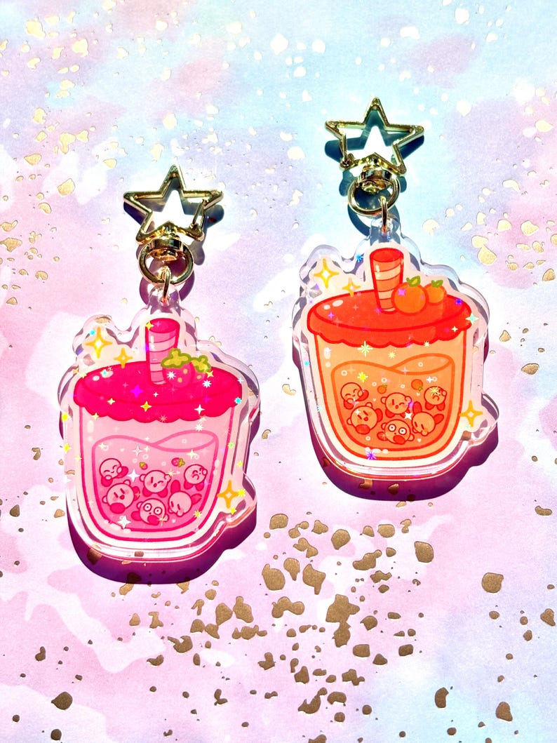 May include: Two acrylic keychains in the shape of bubble tea drinks. One is pink with a strawberry topping, the other is orange with a peach topping. Both have gold star-shaped clasps and feature cartoon characters.