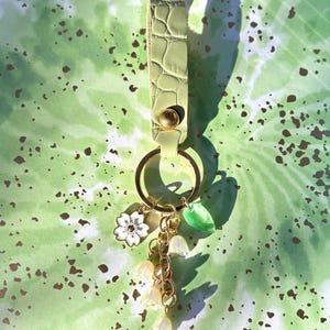 May include: A light green leather keychain with gold-colored hardware. The keychain features a gold ring, a white flower charm, a green leaf charm, and a cluster of small white flower charms hanging from a gold chain.