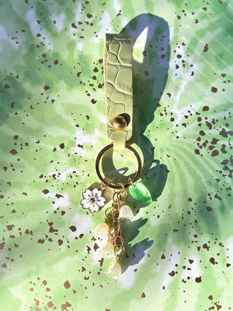 May include: A light green leather keychain with gold-colored hardware. The keychain features a gold ring, a white flower charm, a green leaf charm, and a cluster of small white flower charms hanging from a gold chain.
