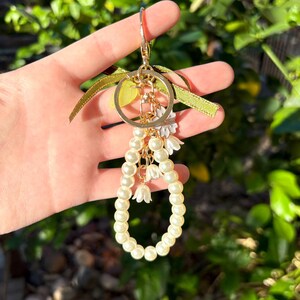 May include: A gold-colored keychain with a large ring, adorned with a cluster of faux pearls and small white flower charms. The keychain also features a green ribbon and a small gold-colored charm. The background is a blurred view of green foliage.