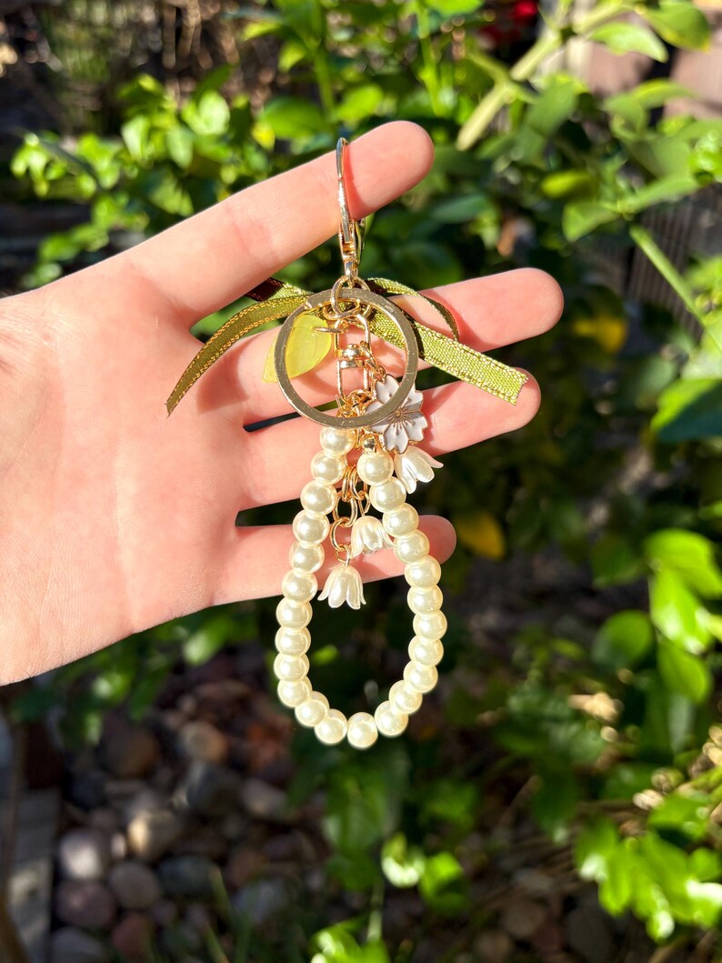May include: A gold-colored keychain with a large ring, adorned with a cluster of faux pearls and small white flower charms. The keychain also features a green ribbon and a small gold-colored charm. The background is a blurred view of green foliage.