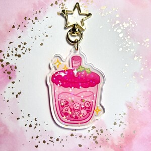 May include: A clear acrylic keychain depicting a pink drink with Kirby characters. The drink has a pink lid with strawberries and sparkles. The keychain has a gold star-shaped clasp.