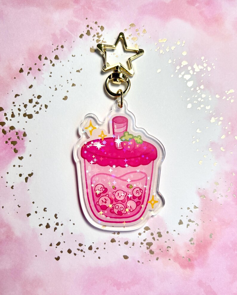 May include: A clear acrylic keychain depicting a pink drink with Kirby characters. The drink has a pink lid with strawberries and sparkles. The keychain has a gold star-shaped clasp.