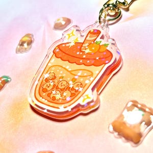 May include: A clear acrylic bubble tea keychain with orange and yellow details. The design includes cartoon characters within the drink, and a gold star-shaped clasp. The background is a soft pink. The keychain is a fun accessory.