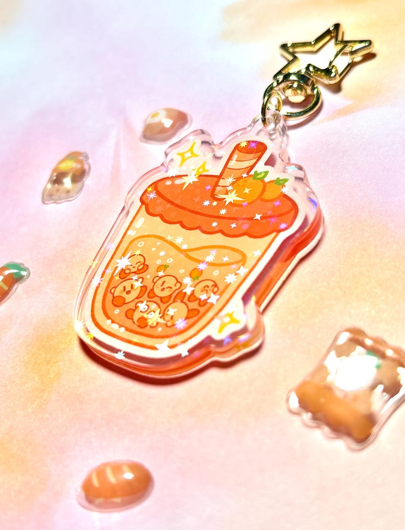 May include: A clear acrylic bubble tea keychain with orange and yellow details. The design includes cartoon characters within the drink, and a gold star-shaped clasp. The background is a soft pink. The keychain is a fun accessory.