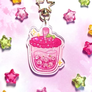 May include: A pink acrylic keychain depicting a cartoon drink with a strawberry and small characters inside. The keychain has a gold star-shaped clasp and is surrounded by star-shaped beads in various colours, set against a pink background.