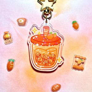 May include: A clear acrylic keychain with a cartoon drink design in orange and yellow, a gold star clasp, and food-themed charms. The drink features orange fruit and cartoon characters. Small food charms surround the keychain. The keychain is approximately 8 cm in length.