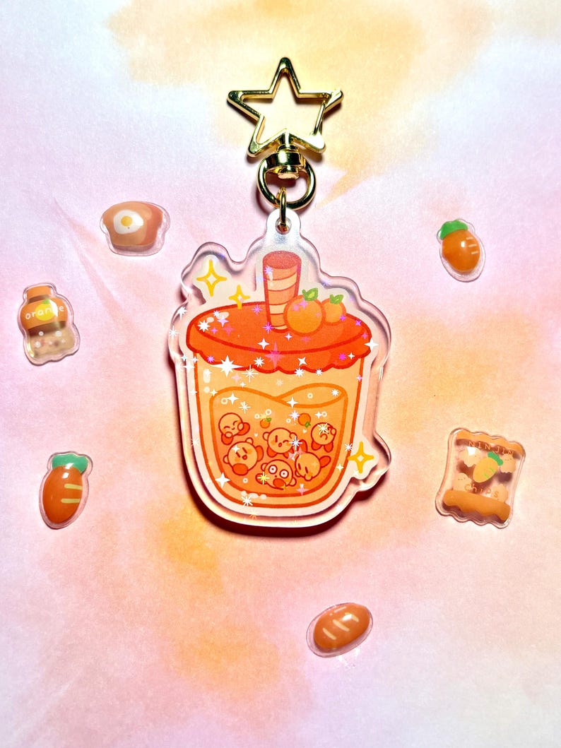 May include: A clear acrylic keychain with a cartoon drink design in orange and yellow, a gold star clasp, and food-themed charms. The drink features orange fruit and cartoon characters. Small food charms surround the keychain. The keychain is approximately 8 cm in length.