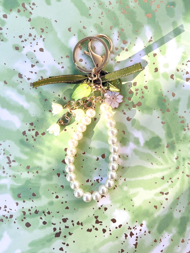 May include: A gold-colored keychain with a pearl beaded loop. The keychain has a green ribbon, small white flower, and leaf charms. The background is a green and white watercolor design with brown speckles.