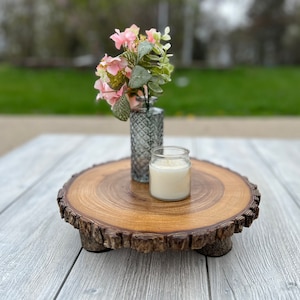 May include: A wooden slice with bark edges, serving as a decorative tray. It features a small white candle and a glass vase with pink flowers.