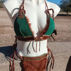 May include: A green and brown leather and fur bikini top and bottom with intricate floral designs. The top has a halter style with a braided strap and the bottom has a high-waisted design with a fringe.