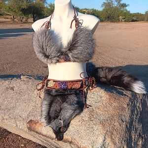 May include: A faux fur bikini set in shades of gray and black, displayed on a mannequin. The set includes a halter top, a belt with floral embroidery, and a matching bottom. A faux fur tail is also included. The set is displayed on a large rock.