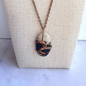 May include: A necklace featuring an oval-shaped stone with a dark blue bottom half and a cream-colored top half. The stone is secured in a decorative copper wire wrap design and hangs from a matching copper-toned metal chain link necklace.