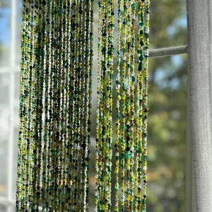 May include: A beaded curtain with strands of green, yellow, and brown beads. The beads vary in size and shape, creating a textured effect. The curtain hangs in front of a window, with a white curtain visible on the right side.