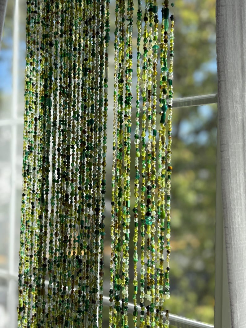 May include: A beaded curtain with strands of green, yellow, and brown beads. The beads vary in size and shape, creating a textured effect. The curtain hangs in front of a window, with a white curtain visible on the right side.