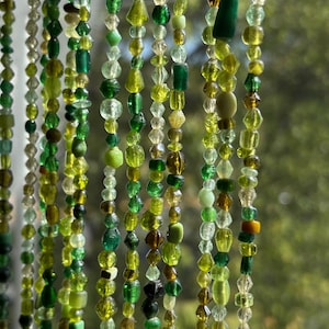 May include: Strands of green beads in various shapes and sizes hang vertically. The beads range in color from light yellow-green to deep forest green, with some clear and brown beads interspersed. The beads appear to be strung together, creating a decorative element.