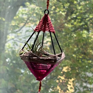 May include: A pink and brown macrame plant hanger with a small air plant inside. The hanger is suspended from a window with a pink crystal hanging from the bottom.