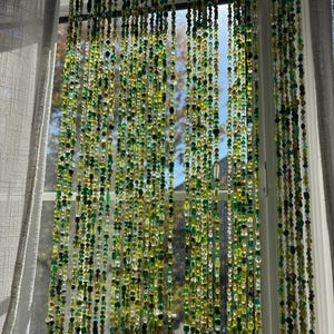 A beaded curtain hangs in front of a window, featuring strands of green and yellow beads. The beads vary in size and shape, creating a textured, decorative effect. The curtain allows light to filter through, with a view of the outside visible.