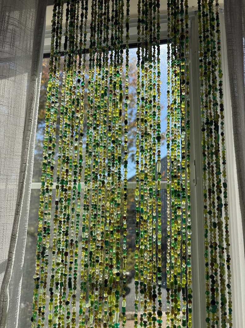 A beaded curtain hangs in front of a window, featuring strands of green and yellow beads. The beads vary in size and shape, creating a textured, decorative effect. The curtain allows light to filter through, with a view of the outside visible.