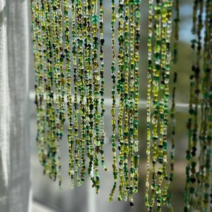 May include: Strands of green beads hang in front of a window. The beads vary in shades of green, from light to dark, and are of different shapes and sizes. The beads are strung together, creating a curtain-like effect. The background is blurred, with a glimpse of the outside.