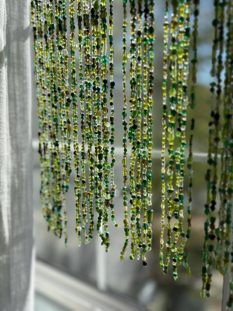 May include: Strands of green beads hang in front of a window. The beads vary in shades of green, from light to dark, and are of different shapes and sizes. The beads are strung together, creating a curtain-like effect. The background is blurred, with a glimpse of the outside.