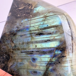 May include: A polished labradorite stone featuring a smooth, reflective surface with prominent iridescent flashes of electric blue and teal. The mineral displays natural linear striations and dark, opaque inclusions throughout its structure.
