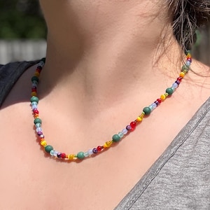 May include: A colorful beaded necklace with a variety of small beads in shades of green, red, yellow, blue, and white. The necklace is strung on a thin cord and is worn around the neck.