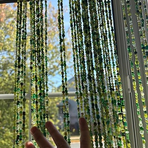 May include: Strands of green and clear beads hang in front of a window. The beads vary in size and shape, creating a textured, decorative curtain. The beads are in shades of green, yellow, and clear, with a hand in the foreground.