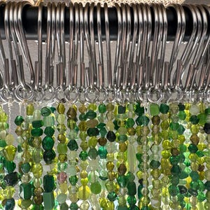 May include: Close-up of a curtain with strands of green and yellow beads hanging from silver metal hooks. The beads are various shapes and sizes, creating a textured and colorful display. The curtain is attached to a black rod.