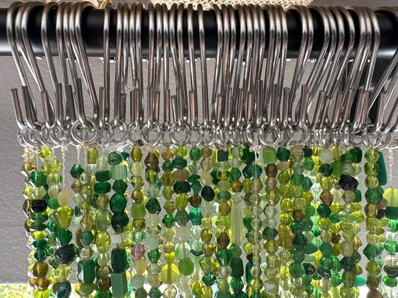 May include: Close-up of a curtain with strands of green and yellow beads hanging from silver metal hooks. The beads are various shapes and sizes, creating a textured and colorful display. The curtain is attached to a black rod.