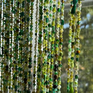 May include: A beaded curtain with strands of various green and clear beads. The beads are different shapes and sizes, creating a textured and layered effect. The curtain hangs in front of a window, with a blurred outdoor view in the background.
