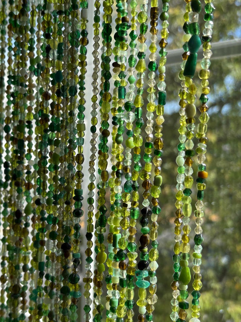 May include: A beaded curtain with strands of various green and clear beads. The beads are different shapes and sizes, creating a textured and layered effect. The curtain hangs in front of a window, with a blurred outdoor view in the background.