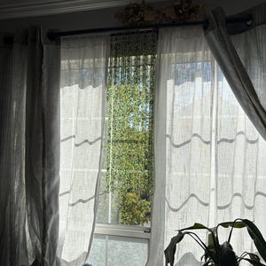 May include: Sheer, light gray curtains hang in front of a window, with sunlight streaming through. The curtains are draped, and a plant is visible in the foreground. A decorative element sits above the window.