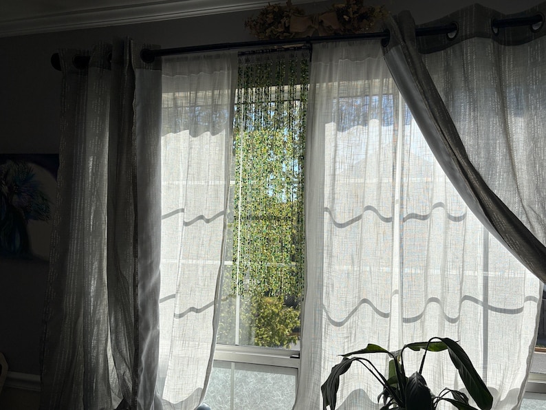 May include: Sheer, light gray curtains hang in front of a window, with sunlight streaming through. The curtains are draped, and a plant is visible in the foreground. A decorative element sits above the window.