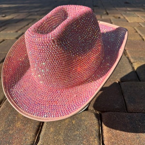 K&ouml;nnte beinhalten: Ein rosa Cowboyhut, der mit schimmernden Strasssteinen besetzt ist. Der Hut liegt auf einem gepflasterten Weg.