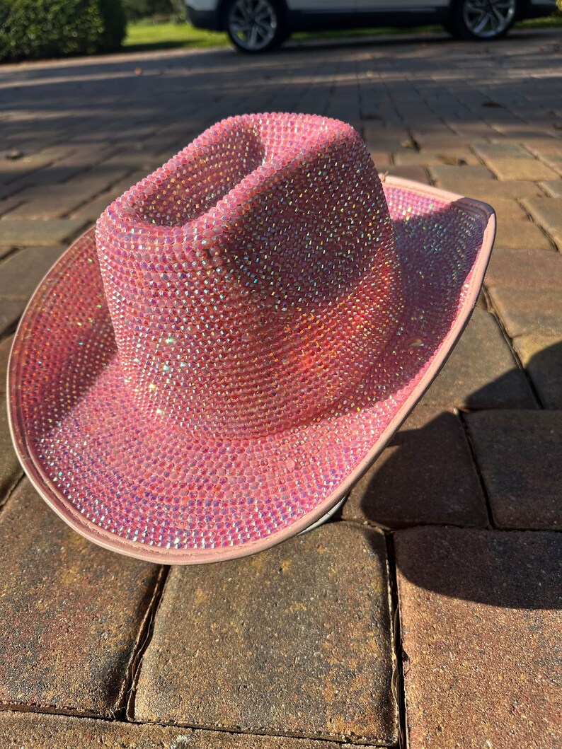 K&ouml;nnte beinhalten: Ein rosa Cowboyhut, der mit schimmernden Strasssteinen besetzt ist. Der Hut liegt auf einem gepflasterten Weg.