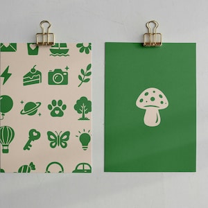 May include: Two rectangular cards held by gold binder clips. The left card is cream-colored with a repeating pattern of green icons including a camera, cake, butterfly, and tree. The right card is solid green with a single cream-colored mushroom icon.
