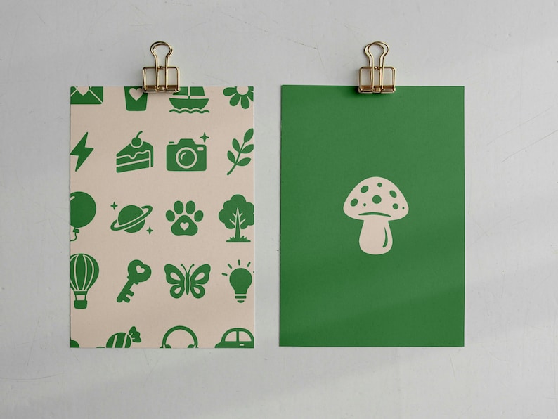May include: Two rectangular cards held by gold binder clips. The left card is cream-colored with a repeating pattern of green icons including a camera, cake, butterfly, and tree. The right card is solid green with a single cream-colored mushroom icon.