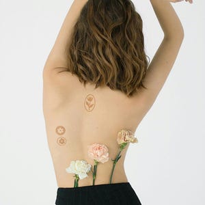 Puede incluir: Un conjunto de tatuajes temporales marrones con un tulip&aacute;n en un marco ovalado y dos peque&ntilde;os dise&ntilde;os florales circulares sobre la piel, acompa&ntilde;ados de tres claveles frescos en blanco, rosa claro y melocot&oacute;n metidos en ropa negra.